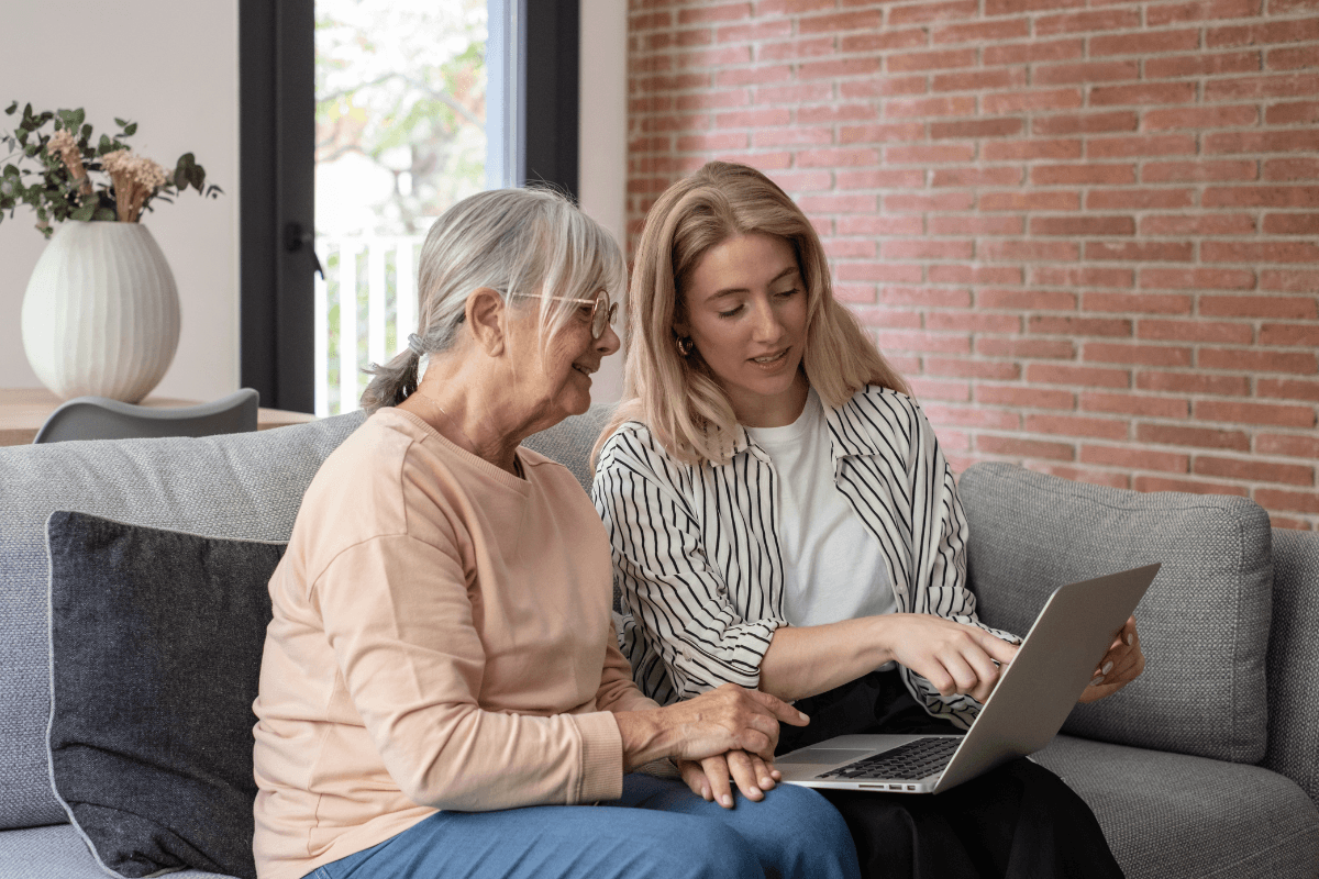Young woman sits with an older family member on a couch, guiding her through a laptop. Warm, supportive interaction highlights learning, technology assistance and multigenerational connection at home.