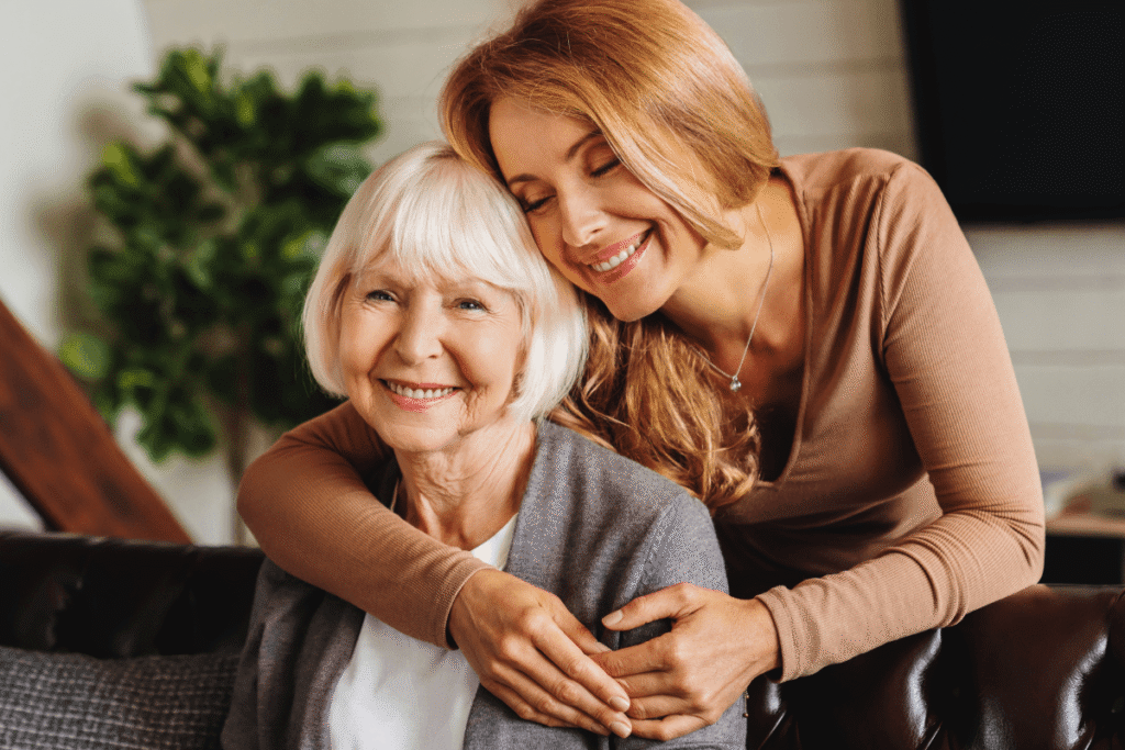 Happy daughter embracing from behind elderly mother at living room. Love and care. Family time and bonding concept. Assistance and support of elder generation.