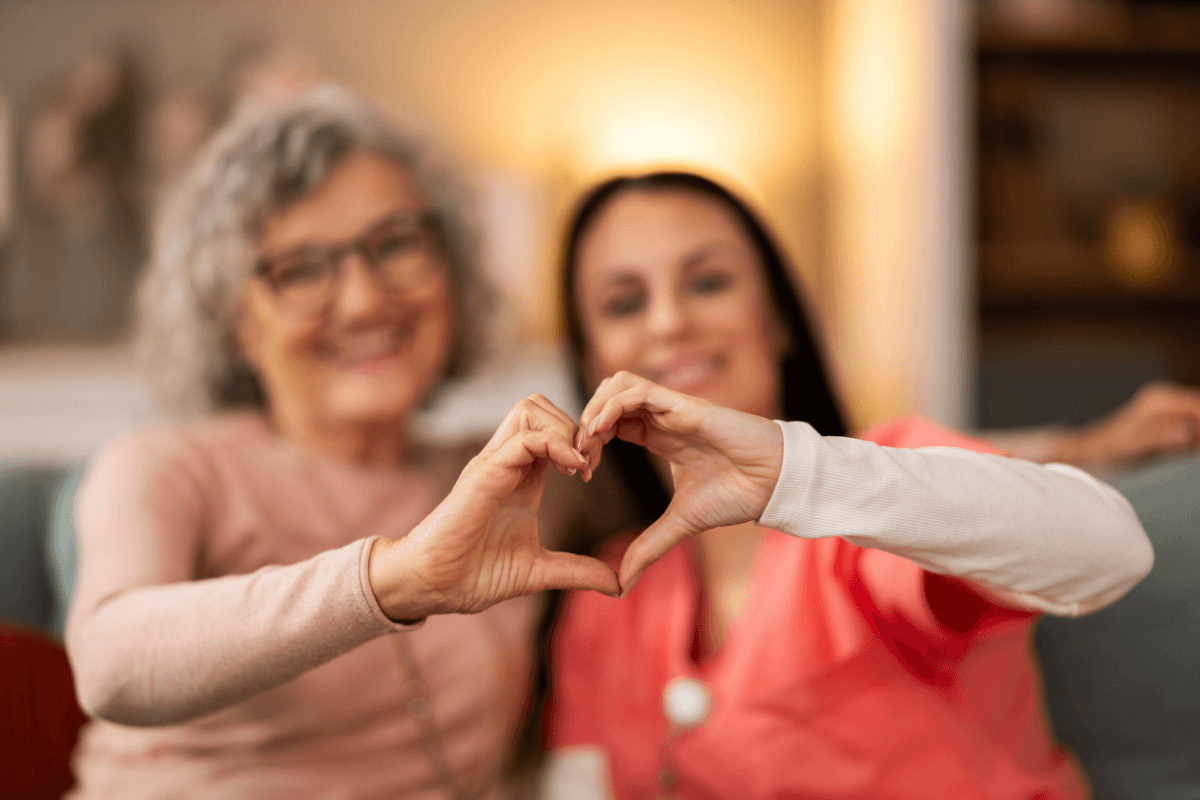Medical worker and senior woman making a heart together with hands while sitting on couch in living room.