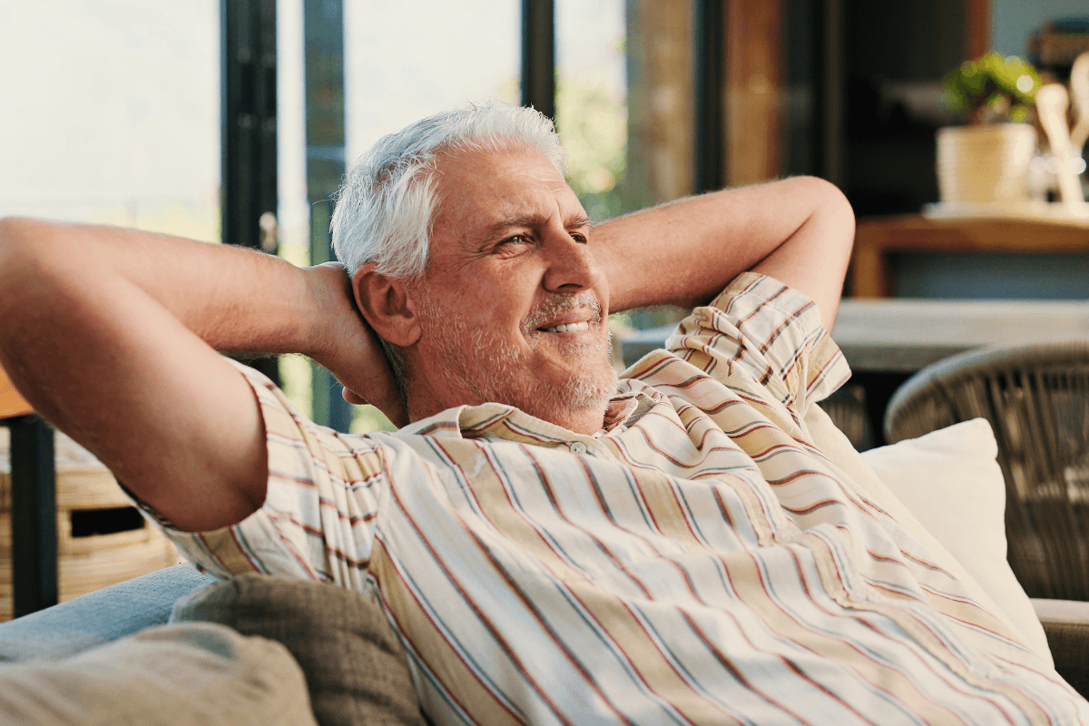 A senior man sits peacefully on the couch, smiling as he enjoys a quiet morning at home.